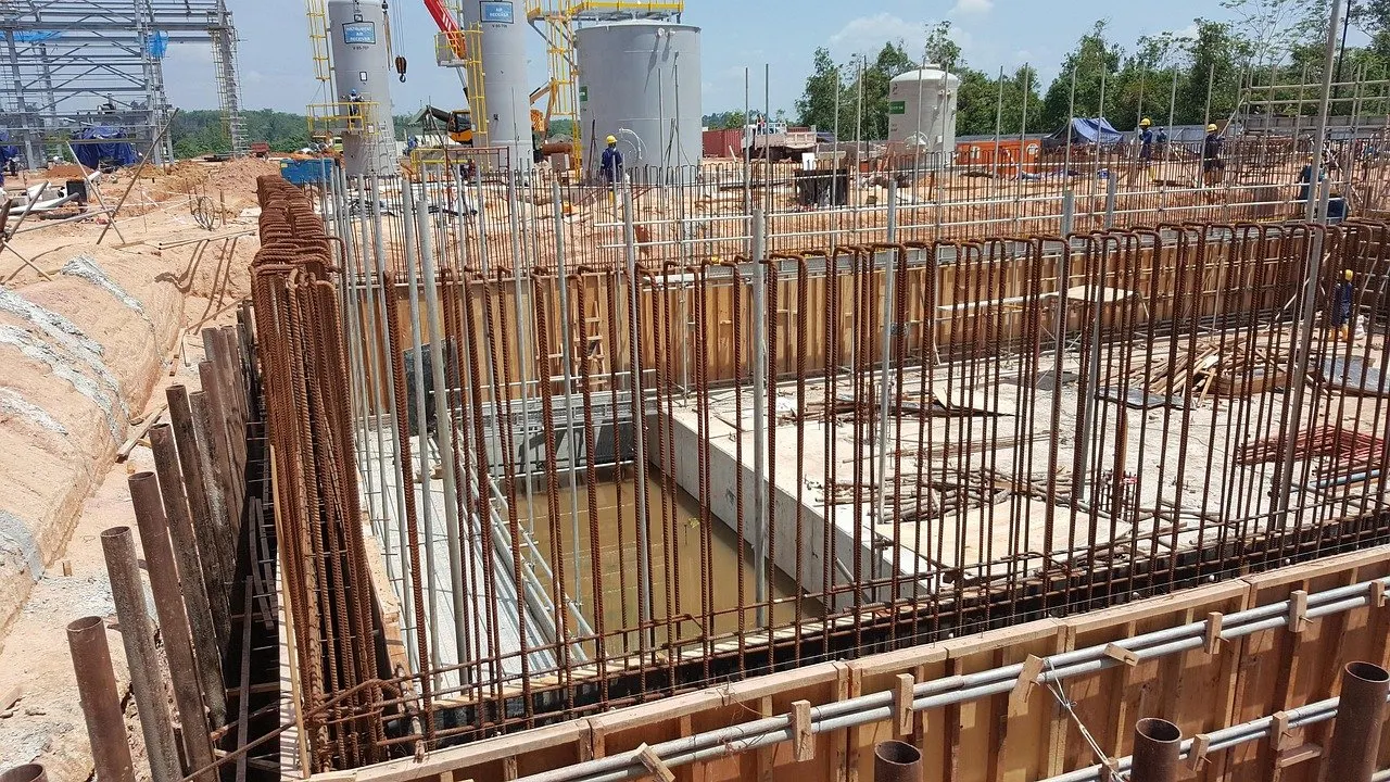 Baugrube mit Bewehrungsstäben und Schalungen, im Hintergrund Arbeiter und Silos.