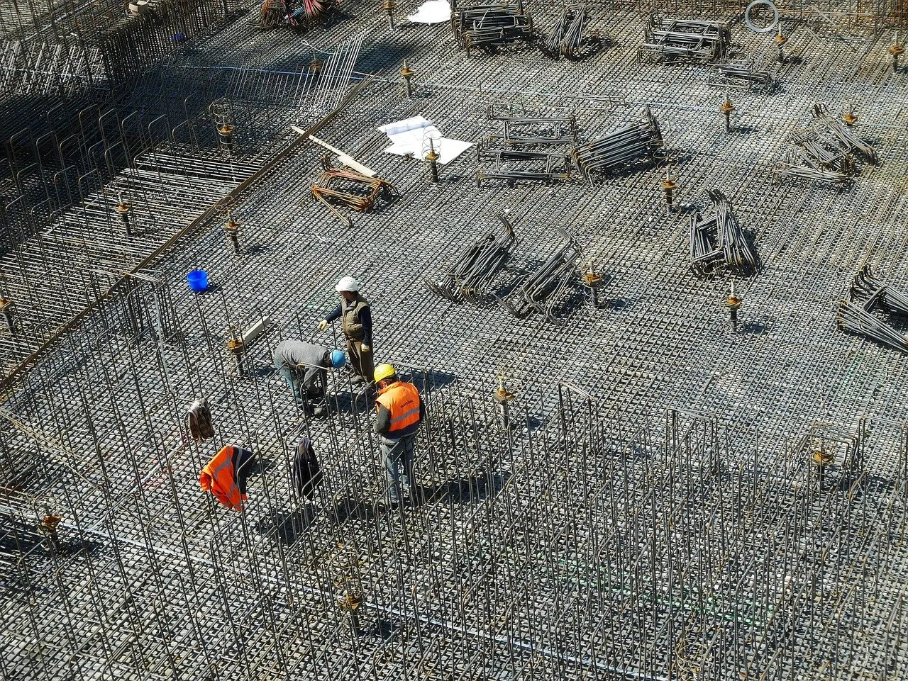 Bewehrungsmatten auf einer Baustelle, Arbeiter mit Helmen und Warnwesten stehen zwischen den Stahlstäben.